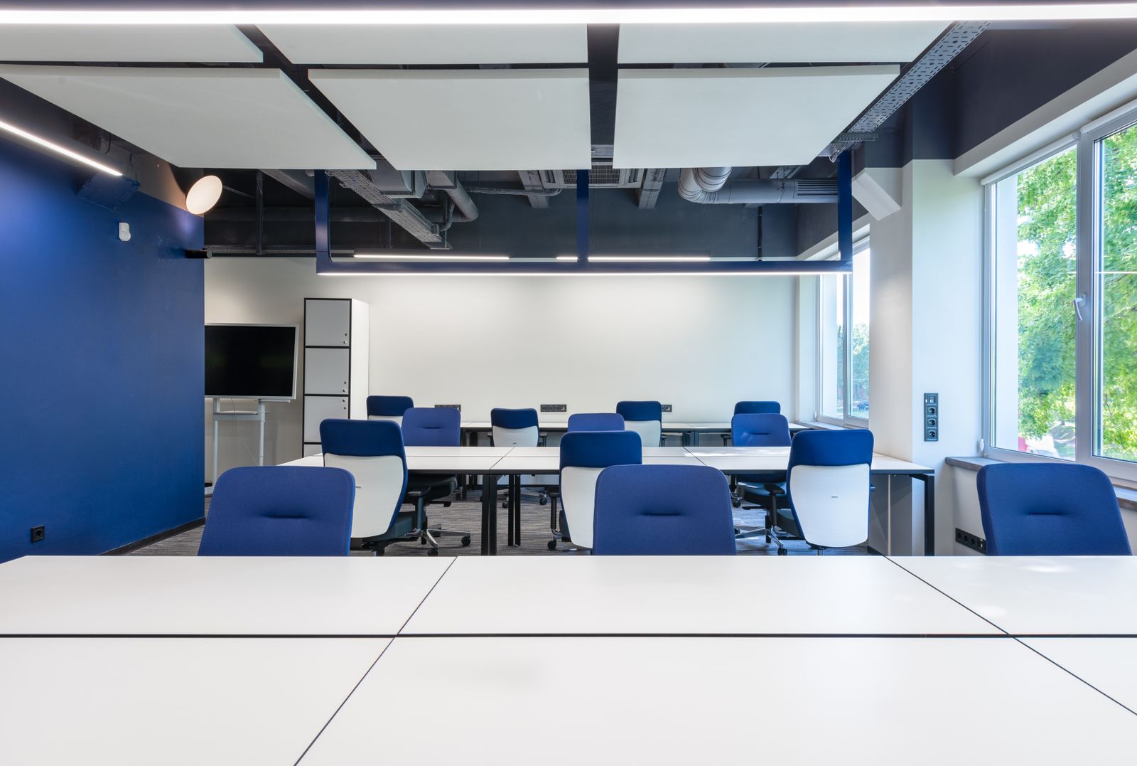 A bright, modern professional office meeting room with white tables, chairs, a wall-mounted display, and large windows