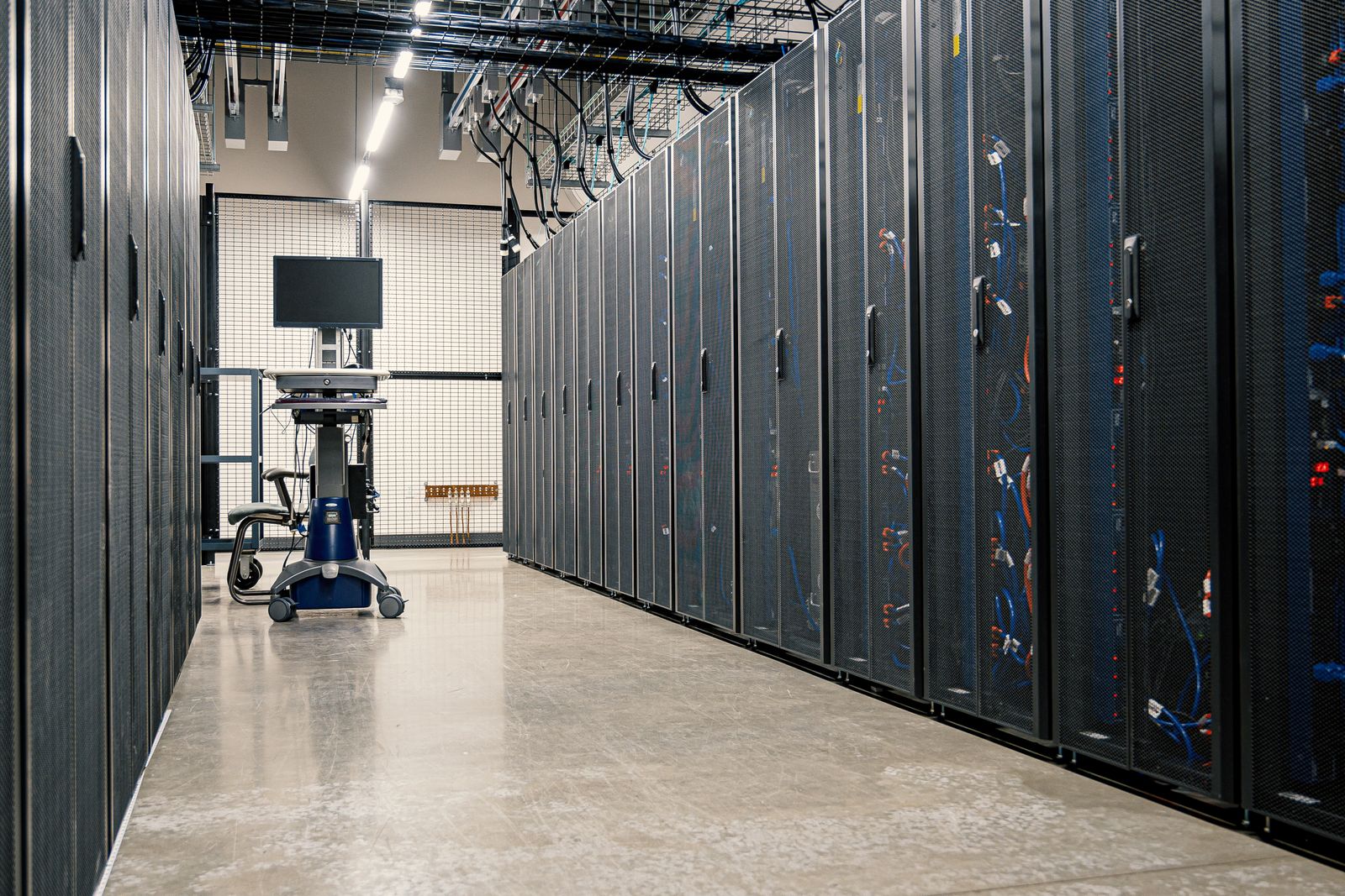 An aisle of black server racks with cabling and a rolling monitor cart in a data center